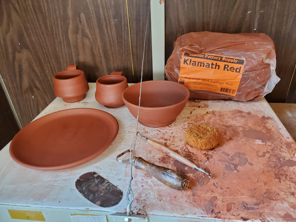 A bowl,  plate, and two mugs made of Seattle Pottery Supply Klamath Red Pottery clay sit on a shelf with pottery tools